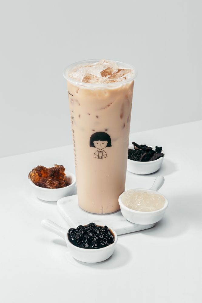 Iced bubble tea in a tall plastic cup with black tapioca pearls, accompanied by small bowls of various toppings including boba, jelly, and syrup, resting on a white surface in Conejo Valley.