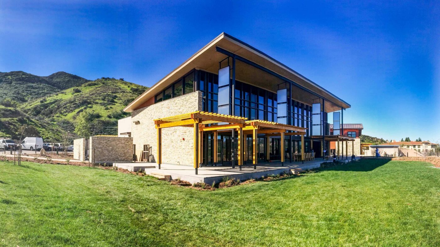 Modern building with large glass windows and wooden pergolas, situated on a lush green lawn.