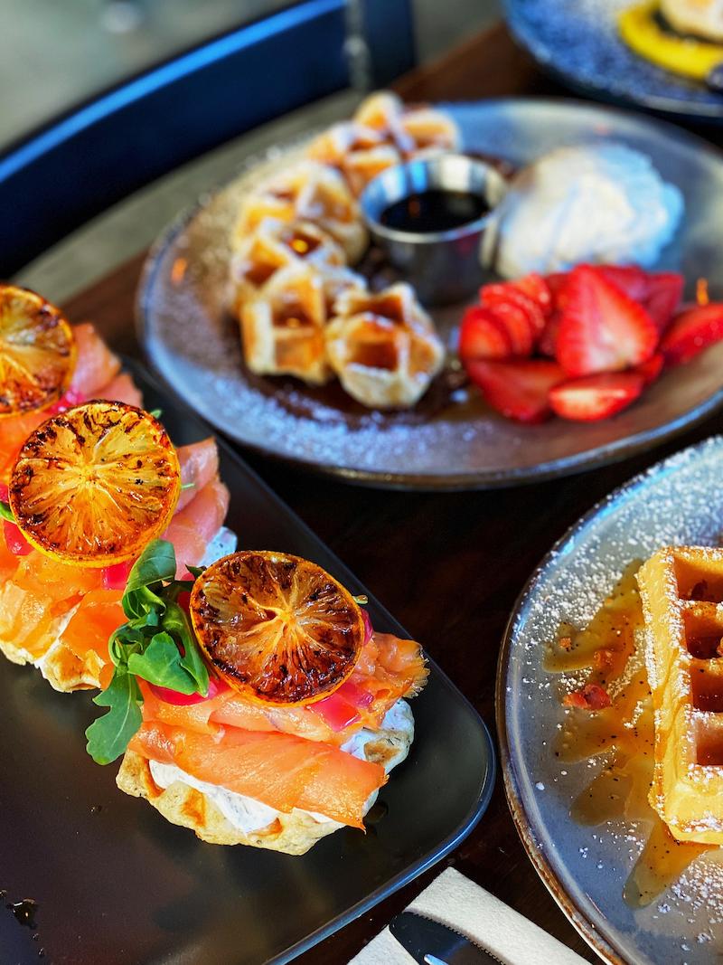 Smoked salmon on waffles topped with grilled orange slices and greens, served on a black plate.