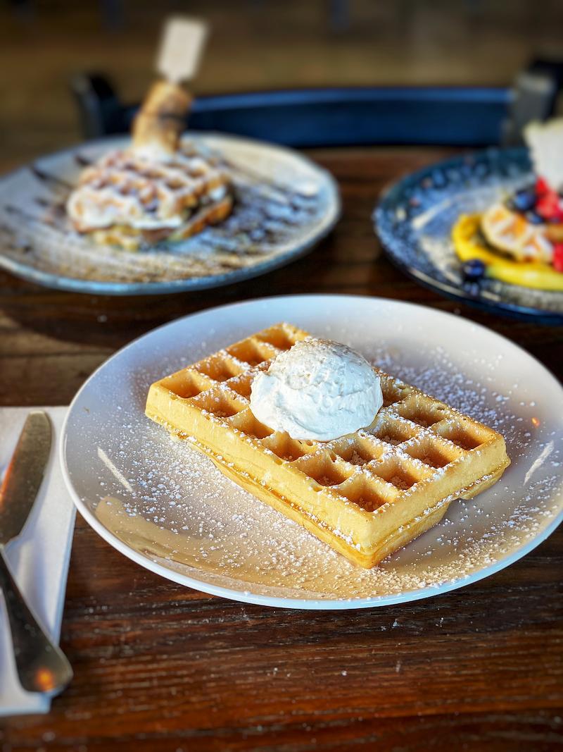Waffle topped with whipped cream, dusted with powdered sugar, on a white plate atop a wooden table.