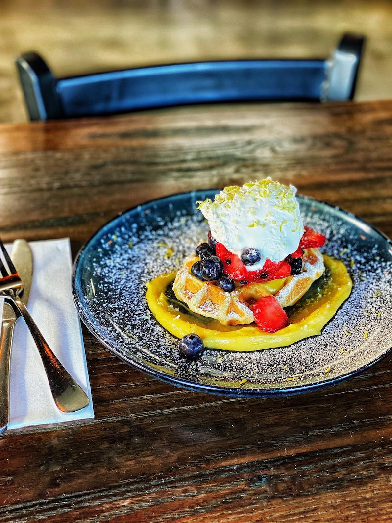 Waffle topped with whipped cream, strawberries, and blueberries, resting on a swirl of mango puree, on a dark plate dusted with powdered sugar, set on a wooden table in Conejo Valley.