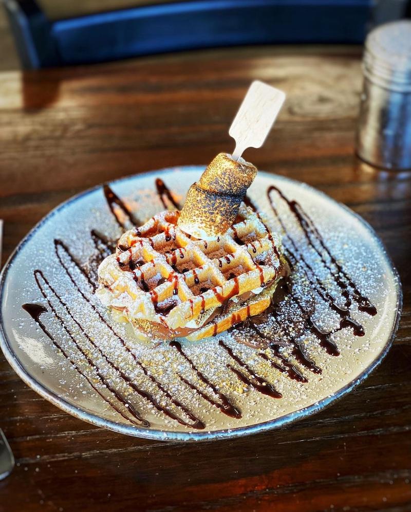 Waffle topped with toasted marshmallow skewer and chocolate drizzle on a powdered sugar-covered plate, set on a wooden table in a café setting in Conejo Valley.