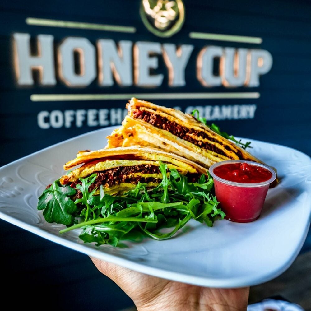 Plate with stacked quesadillas on a bed of arugula, accompanied by red salsa in a cup; held against a wall branded "Honey Cup Coffeehouse & Creamery" in a casual dining setting in Conejo Valley.