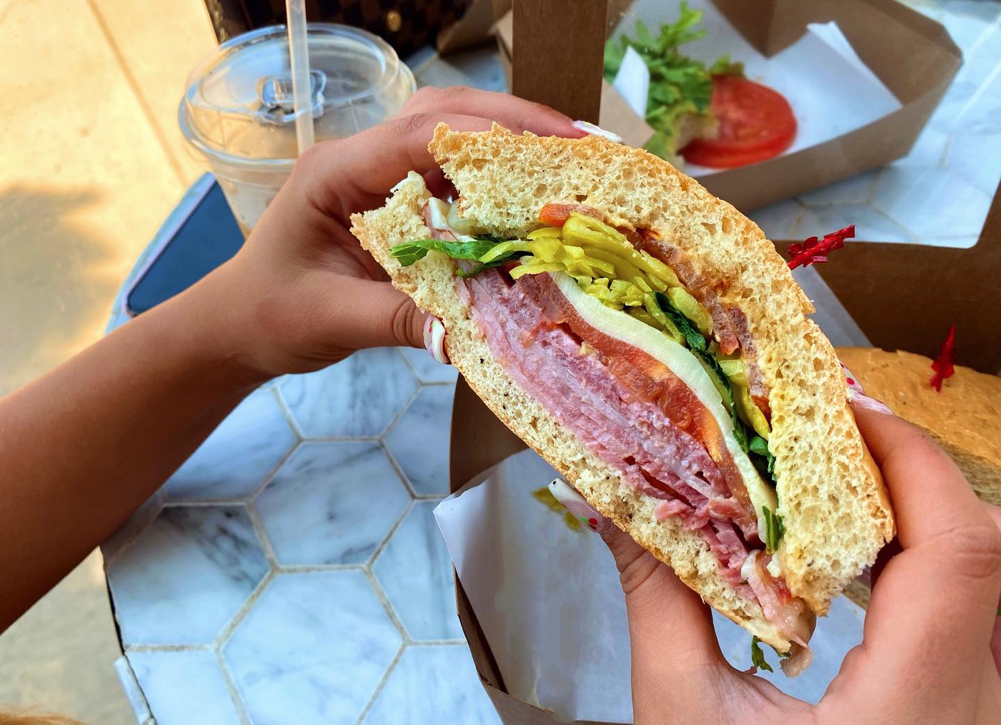 Hands holding a large, stacked sandwich filled with meats, lettuce, and tomato.