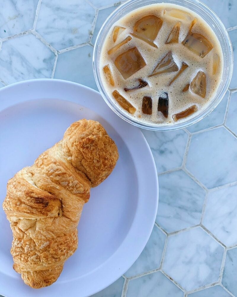 Croissant resting on a white plate beside a cup of iced coffee with visible cubes on a marble hexagonal-tiled surface.