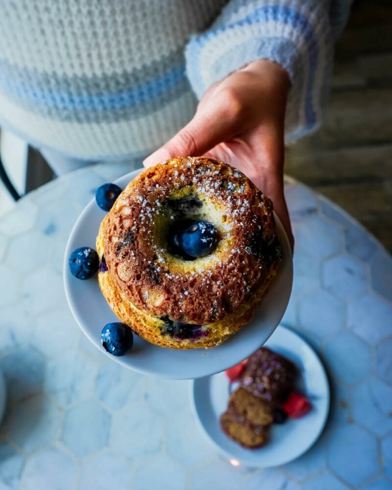 A person holds a blueberry muffin topped with powdered sugar on a small white plate. Surrounding blueberries accent the muffin.