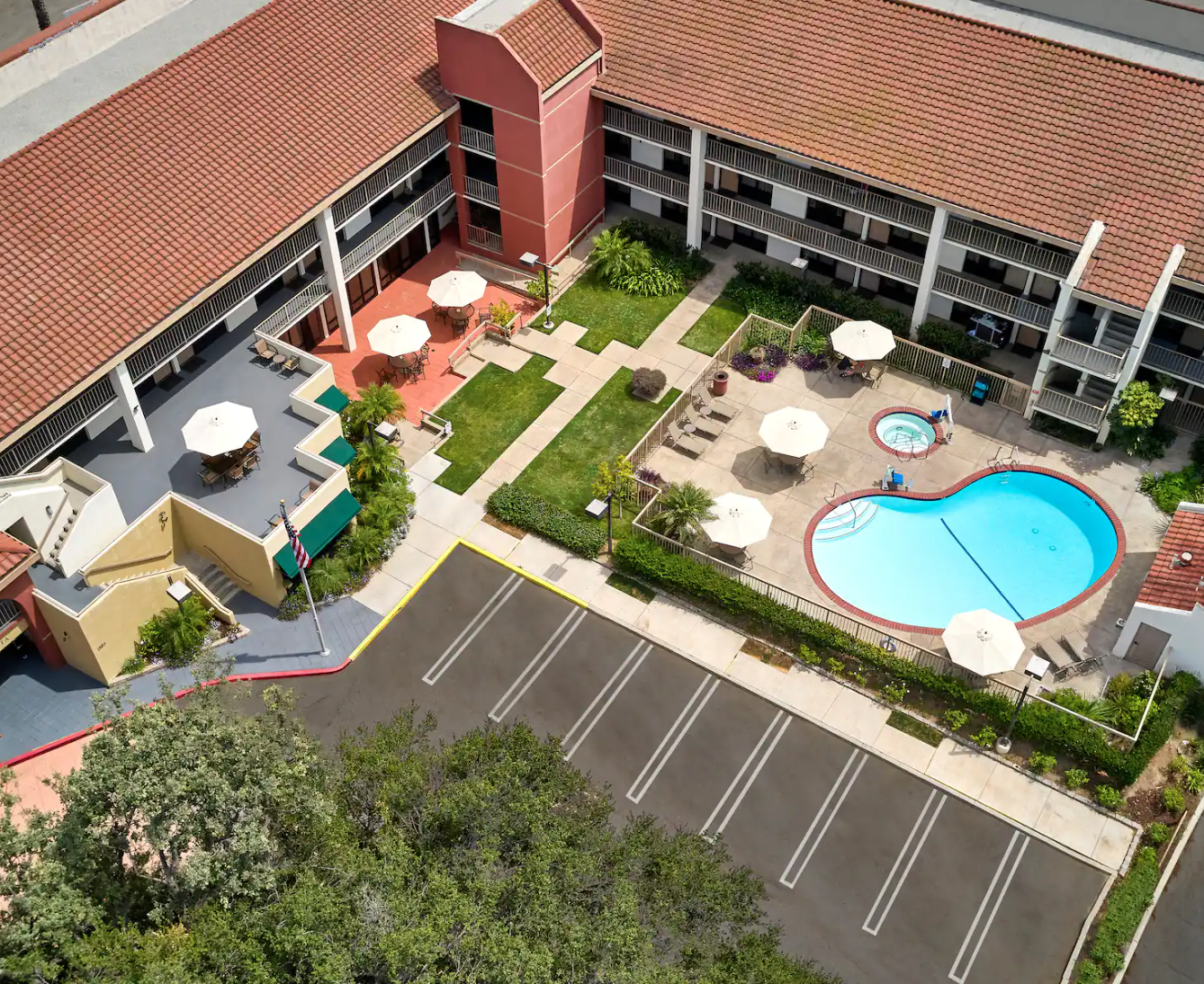 Courtyard pool area with sun umbrellas and lounge chairs surrounded by a path near a multi-story building with red-tiled roofs.