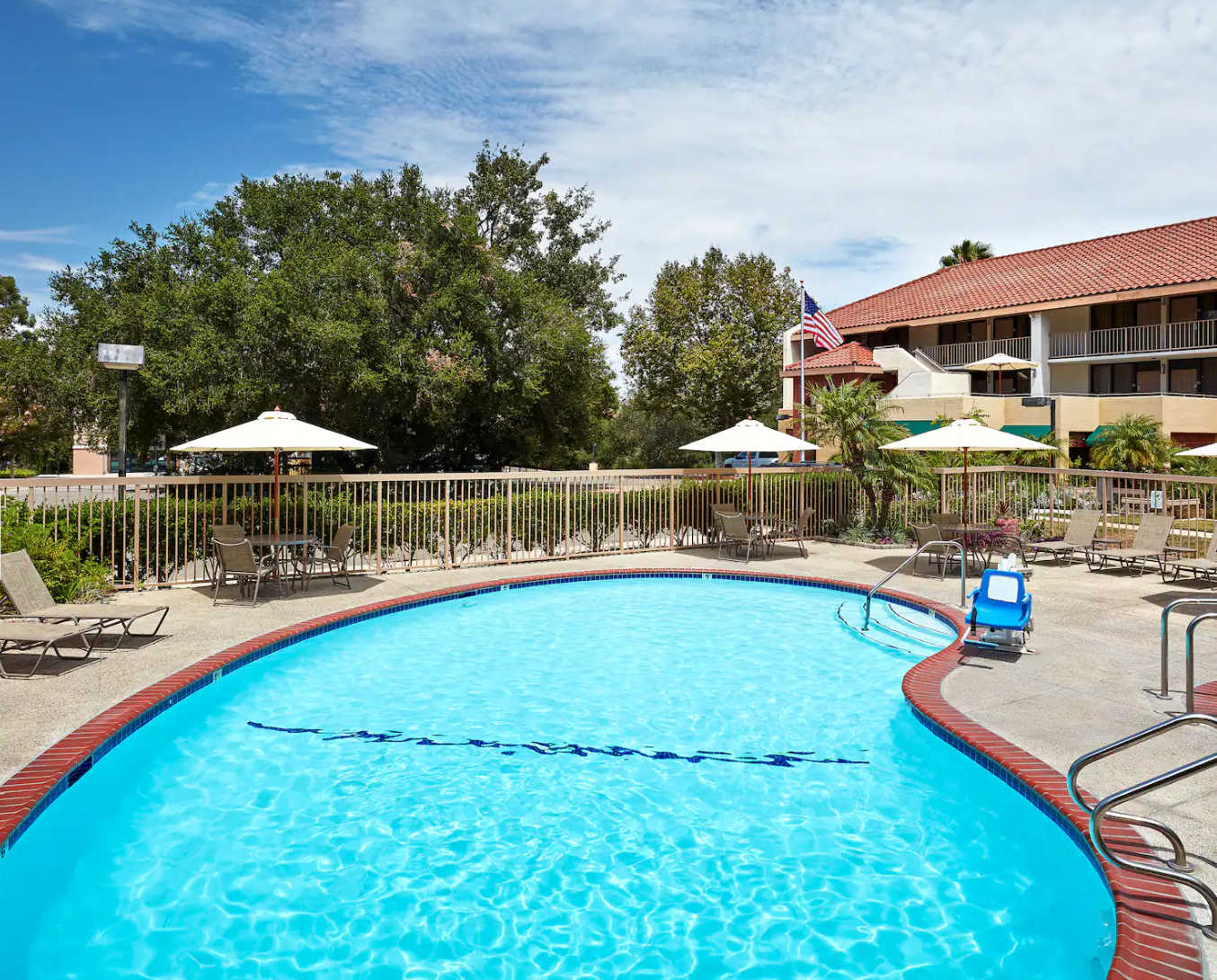 Outdoor oval swimming pool with clear blue water, surrounded by lounge chairs and umbrellas, set in a hotel courtyard with landscaped greenery and a building with a tiled roof in the background in Conejo Valley.