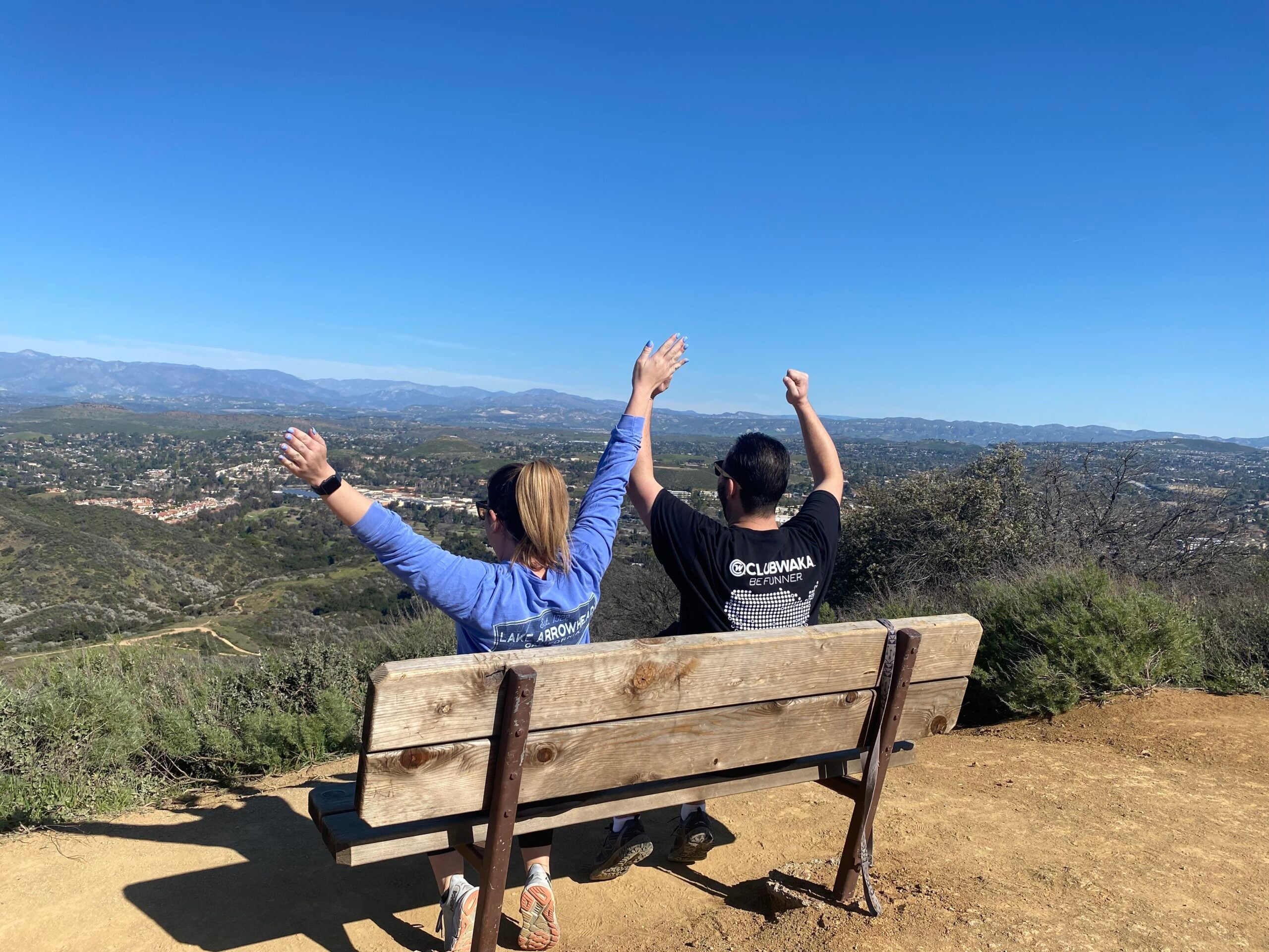 Two people sit on a wooden bench, raising their arms triumphantly, overlooking a vast scenic valley under a clear blue sky.