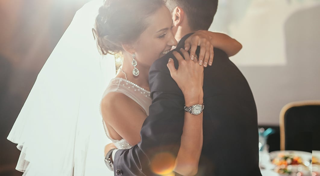 Bride and groom embrace while dancing, illuminated by soft lighting. She wears an elegant veil and earrings, while he sports a watch.