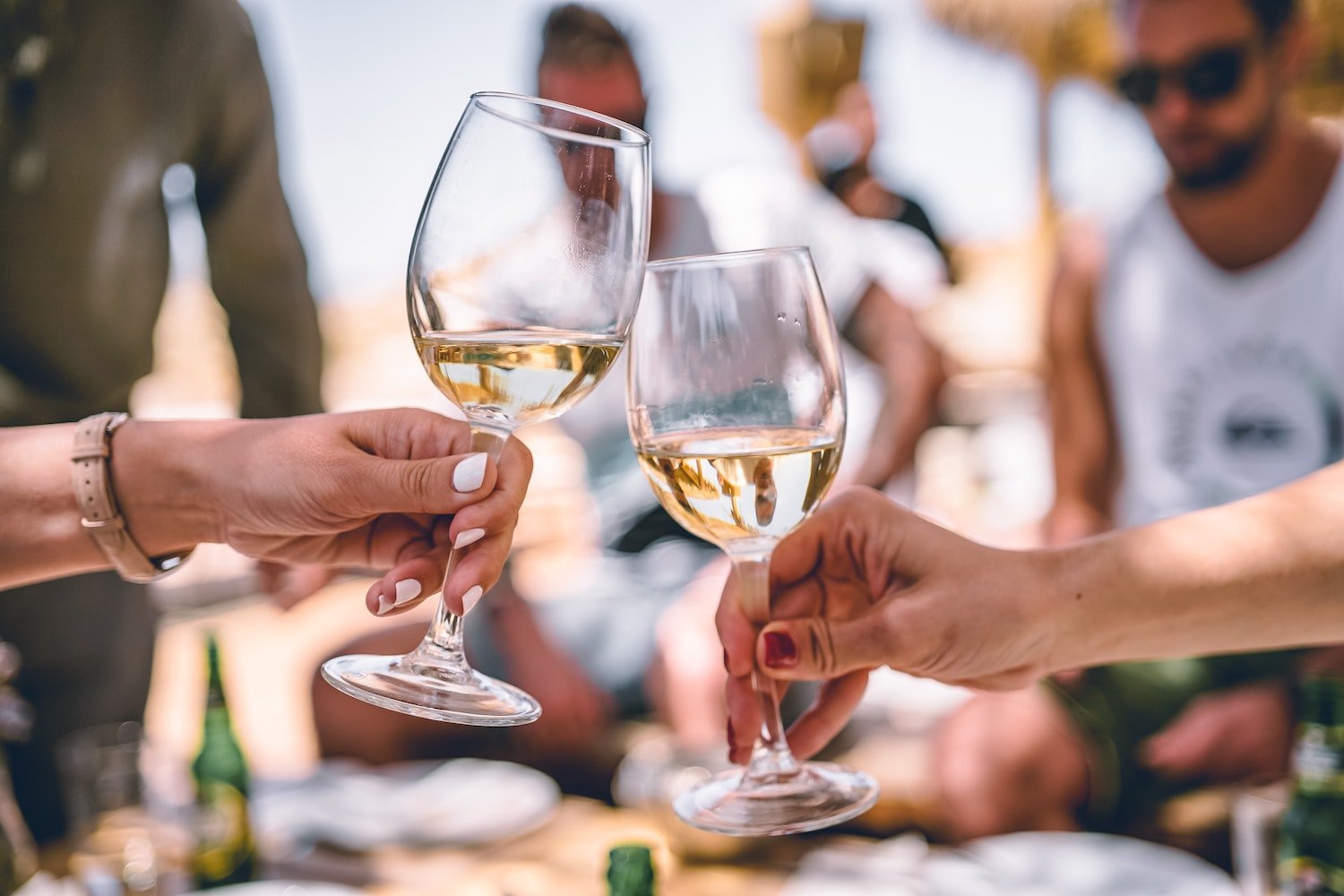 Two hands clinking wine glasses filled with white wine at a social gathering, surrounded by people and sunlit outdoor ambiance with a celebratory and relaxed atmosphere in Conejo Valley.