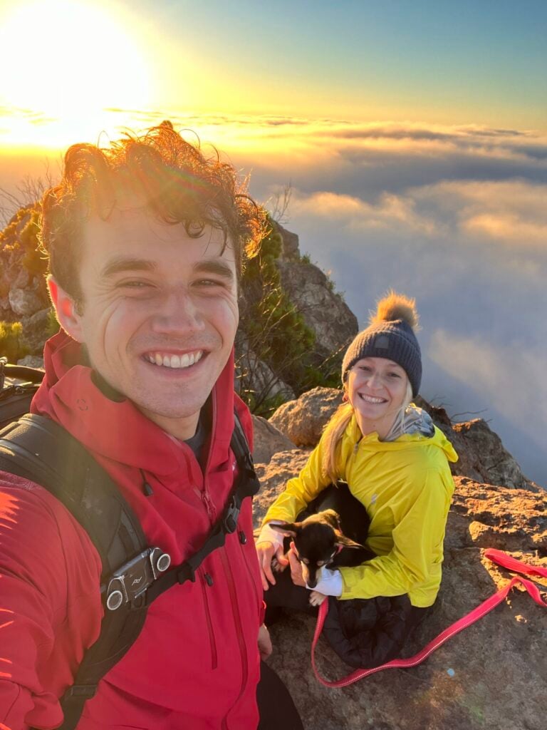 A smiling couple sits on a rocky mountaintop at sunrise. The woman holds a small dog with a pink leash.