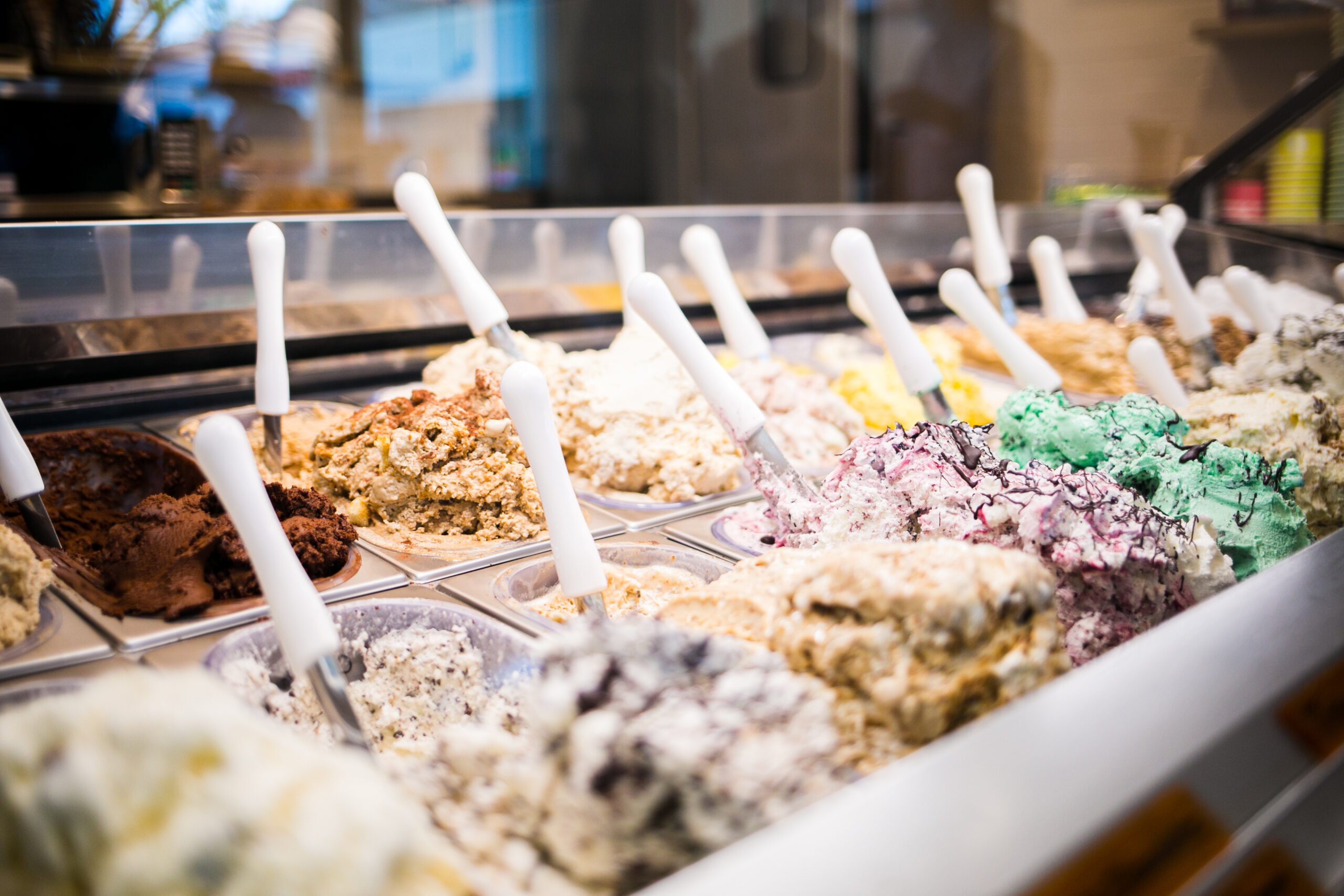 A variety of colorful gelato scoops with white handles are displayed in a freezer case with a tempting dessert selection in a bright ice cream shop setting in Conejo Valley.