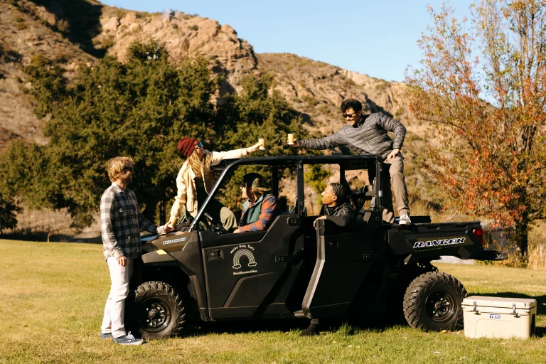 ATV parked on grass with five people interacting cheerfully, surrounded by scenic mountains and trees.