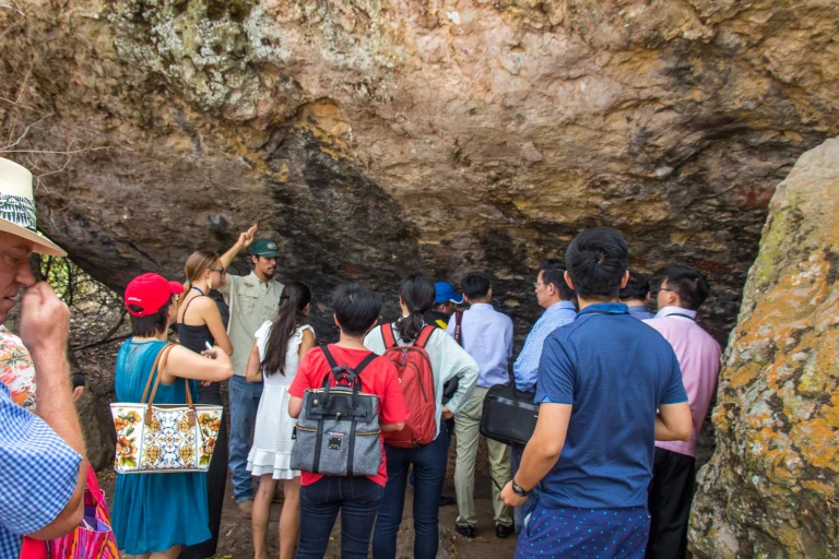 Group of people listening to a guide's explanation near a large, textured rock formation, suggesting a nature tour or educational excursion in a rocky outdoor setting in Conejo Valley wine tasting room.