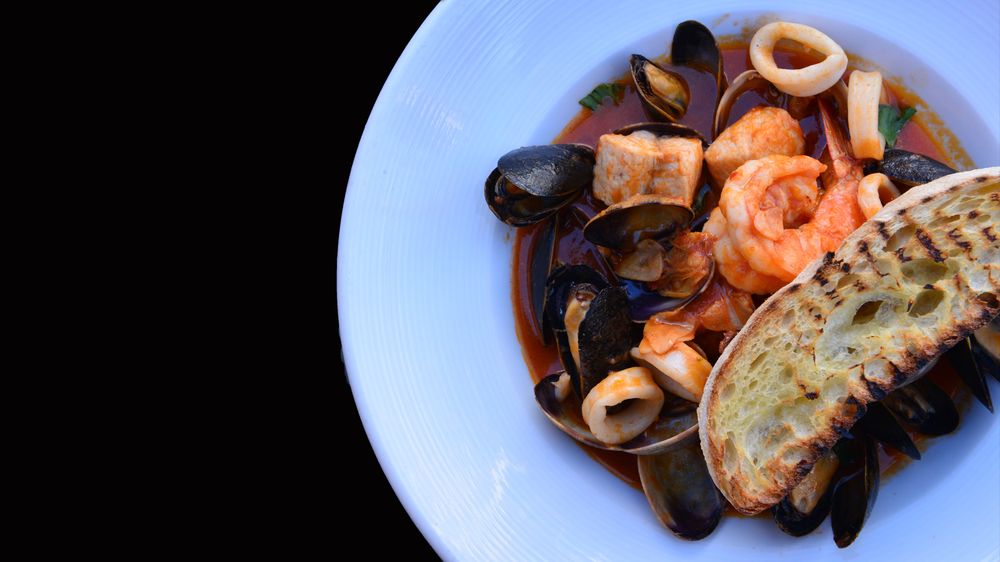 Seafood dish featuring shrimp, mussels, and calamari in a red sauce, topped with a grilled bread slice on a white plate against a black background in Conejo Valley.