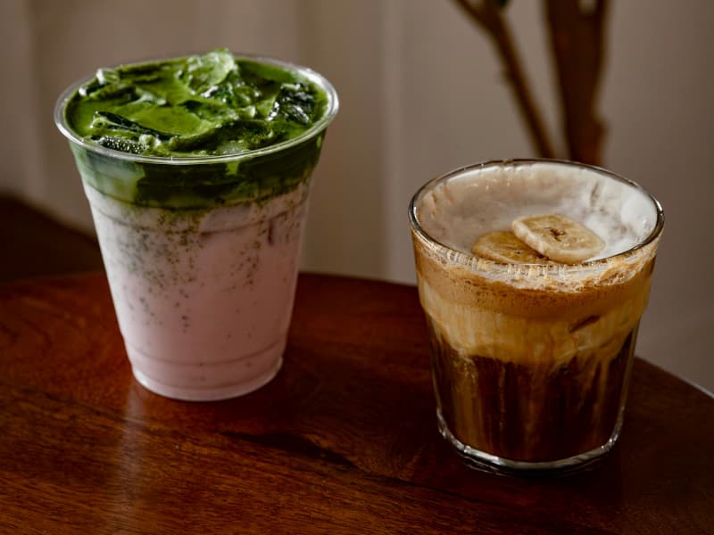A creamy matcha latte and a frothy coffee topped with banana slices sit on a wooden table.