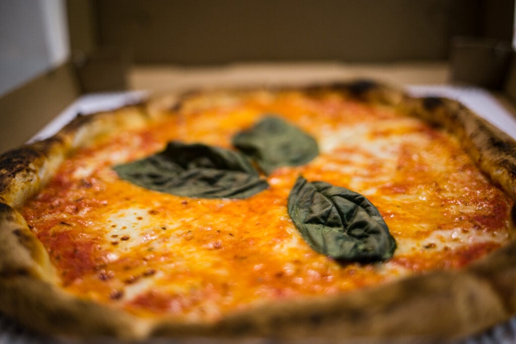 Close-up of a margherita pizza topped with melted cheese, tomato sauce, and several fresh basil leaves in an open takeout box.