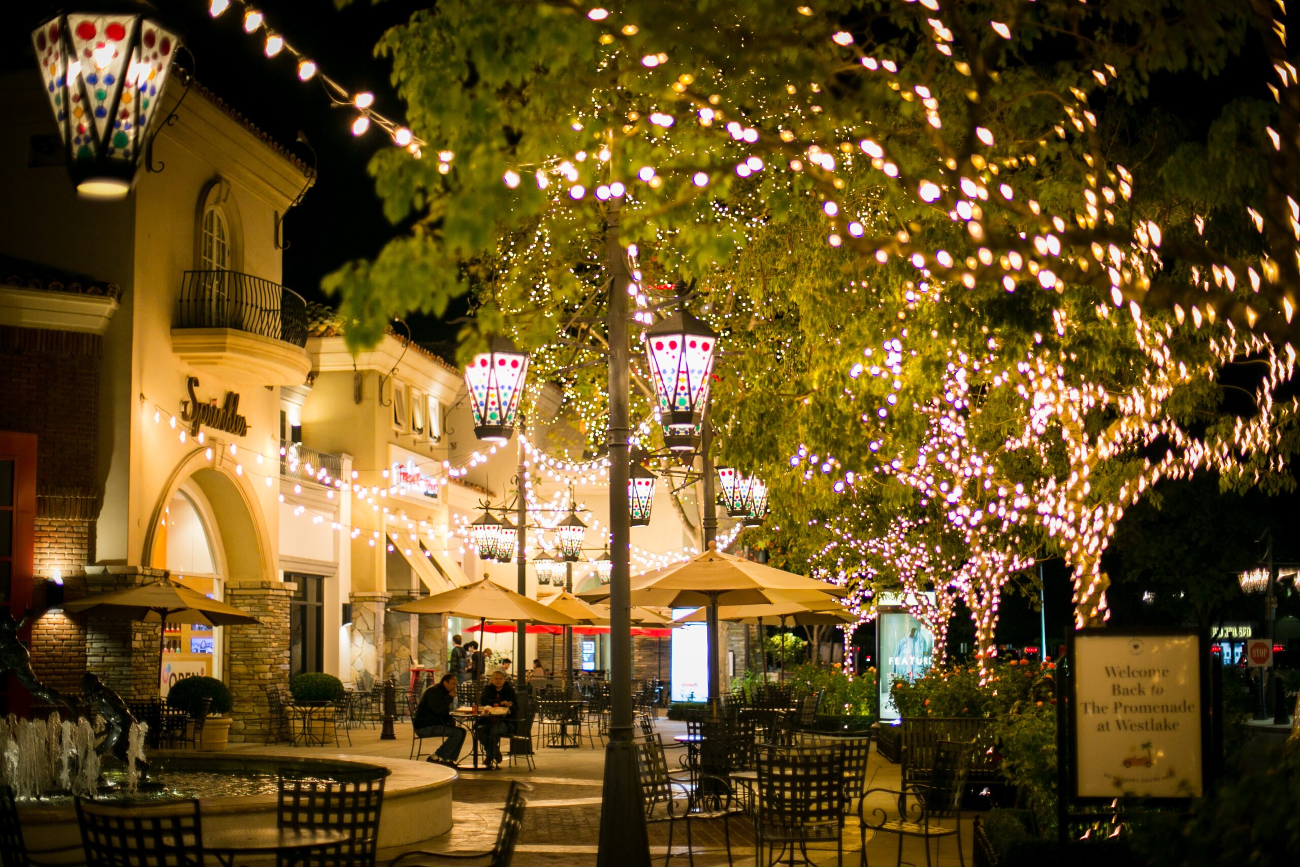 Nighttime scene of an outdoor shopping promenade decorated with string lights and colorful lanterns, with people sitting at café tables under umbrellas and trees wrapped in twinkling lights.