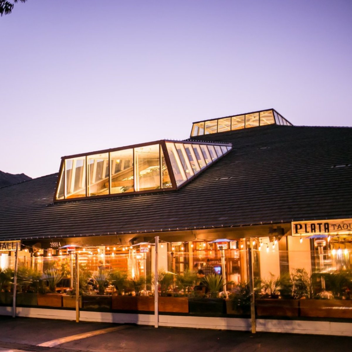 Exterior of Plata Taqueria and Cantina at dusk in Agoura Hills with warm interior lighting, glass skylight windows, and outdoor seating along the front of the restaurant.