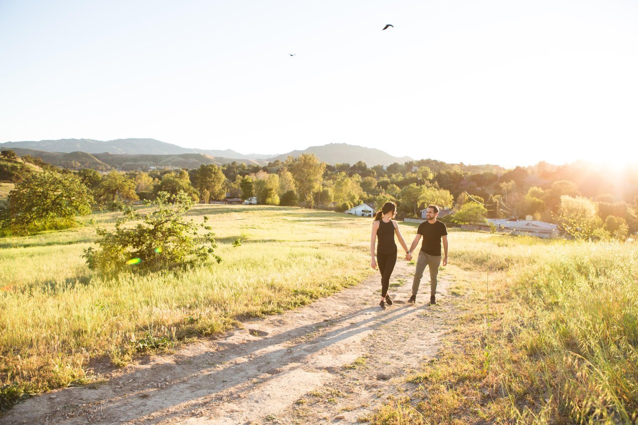 A landscaped park with a winding walking path, green lawns, trees, benches, and a small bridge, with shops and parked cars visible in the background under a cloudy sky.
