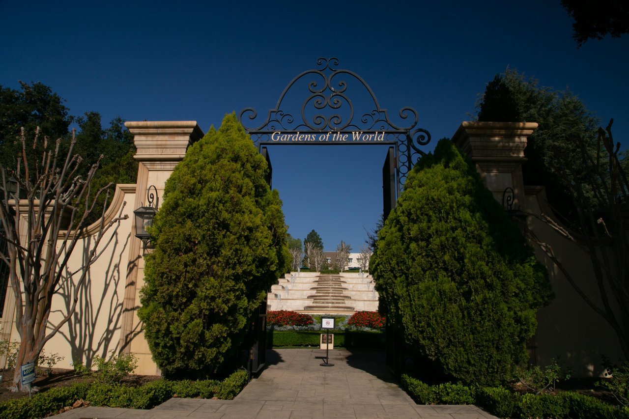 The entrance to Gardens of the World, featuring an ornate iron archway, stone pillars, manicured hedges, and a stairway leading into the garden grounds.