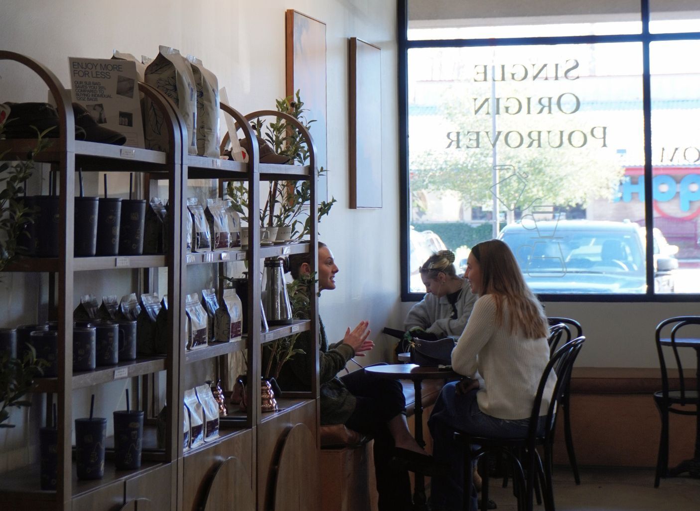 Inside a coffee shop, three people sit and talk at a small table near a large window, with shelves of packaged coffee and merchandise lining the wall.