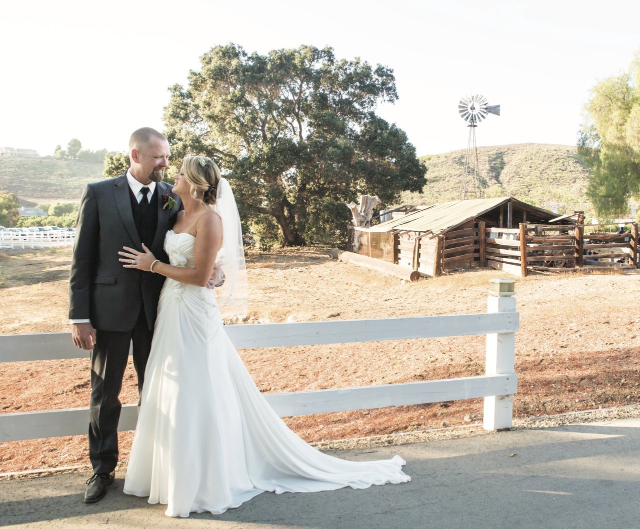A bride and groom stand together by a white fence at a rustic ranch, smiling at each other. The bride wears a white wedding gown and veil, and the groom wears a dark suit. In the background are rolling hills, large trees, a wooden barn structure, and a windmill in soft daylight.