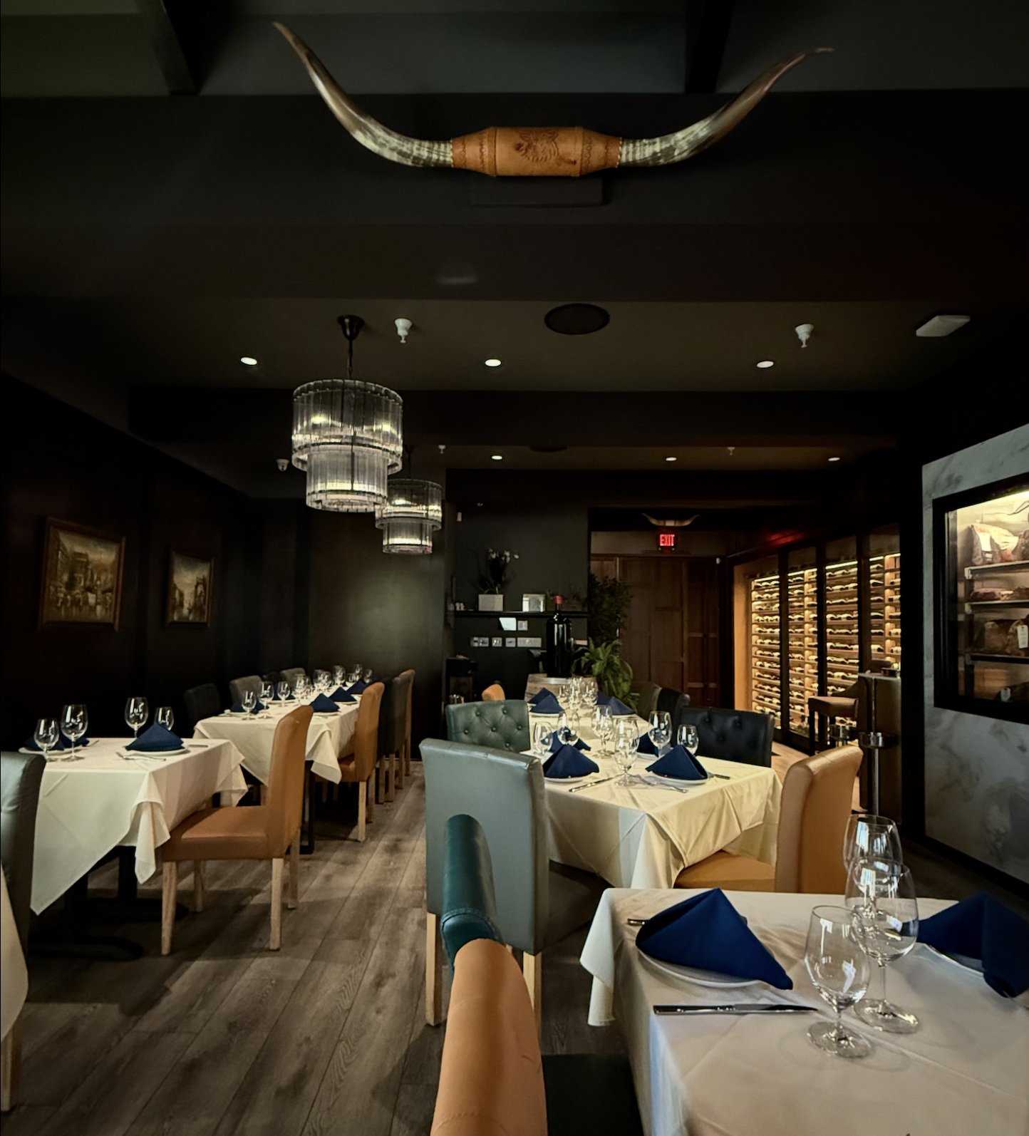 An elegant steakhouse dining room with white tablecloths, neatly set tables, upholstered chairs, modern chandeliers, a wine display, and decorative longhorn horns mounted above.