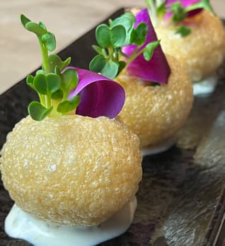 A close-up of crispy, golden-brown appetizer bites served on a dark stone platter, each topped with microgreens and purple edible flower petals.