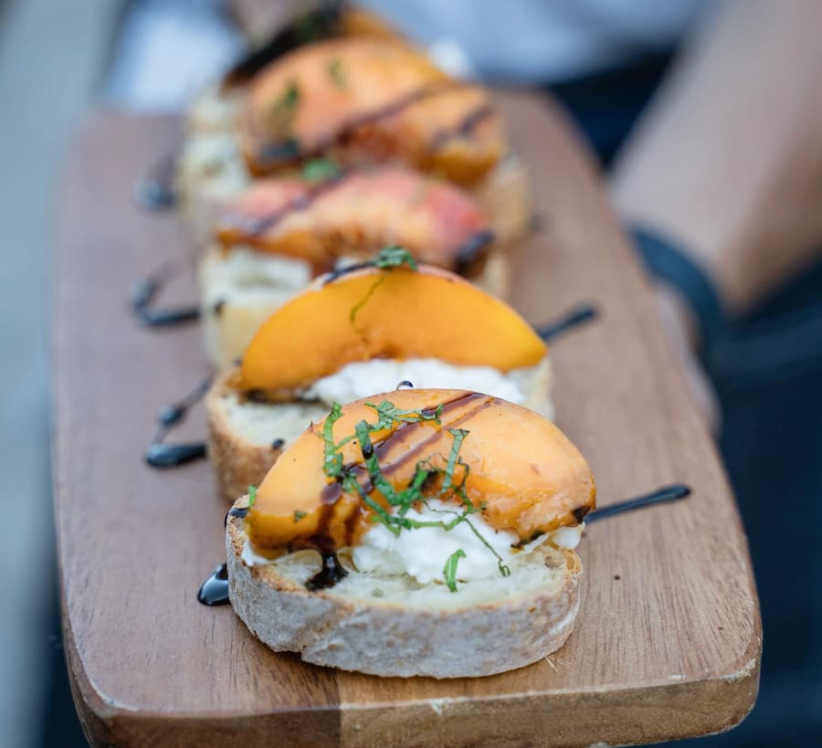 A close-up of dessert crostini served on a wooden board, topped with fresh peach slices, creamy cheese, balsamic drizzle, and chopped herbs.
