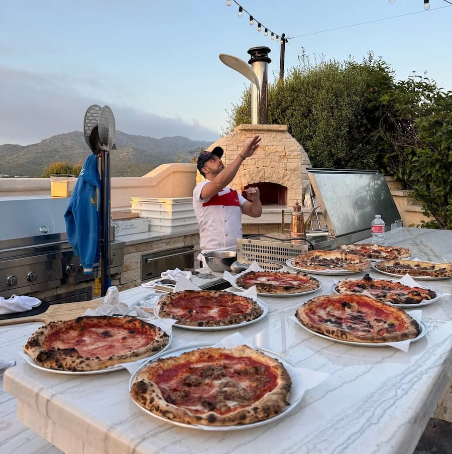 An outdoor pizza station with several freshly baked pizzas displayed on a marble countertop. A chef tosses pizza dough in the air in front of a stone pizza oven, with string lights and rolling hills in the background.