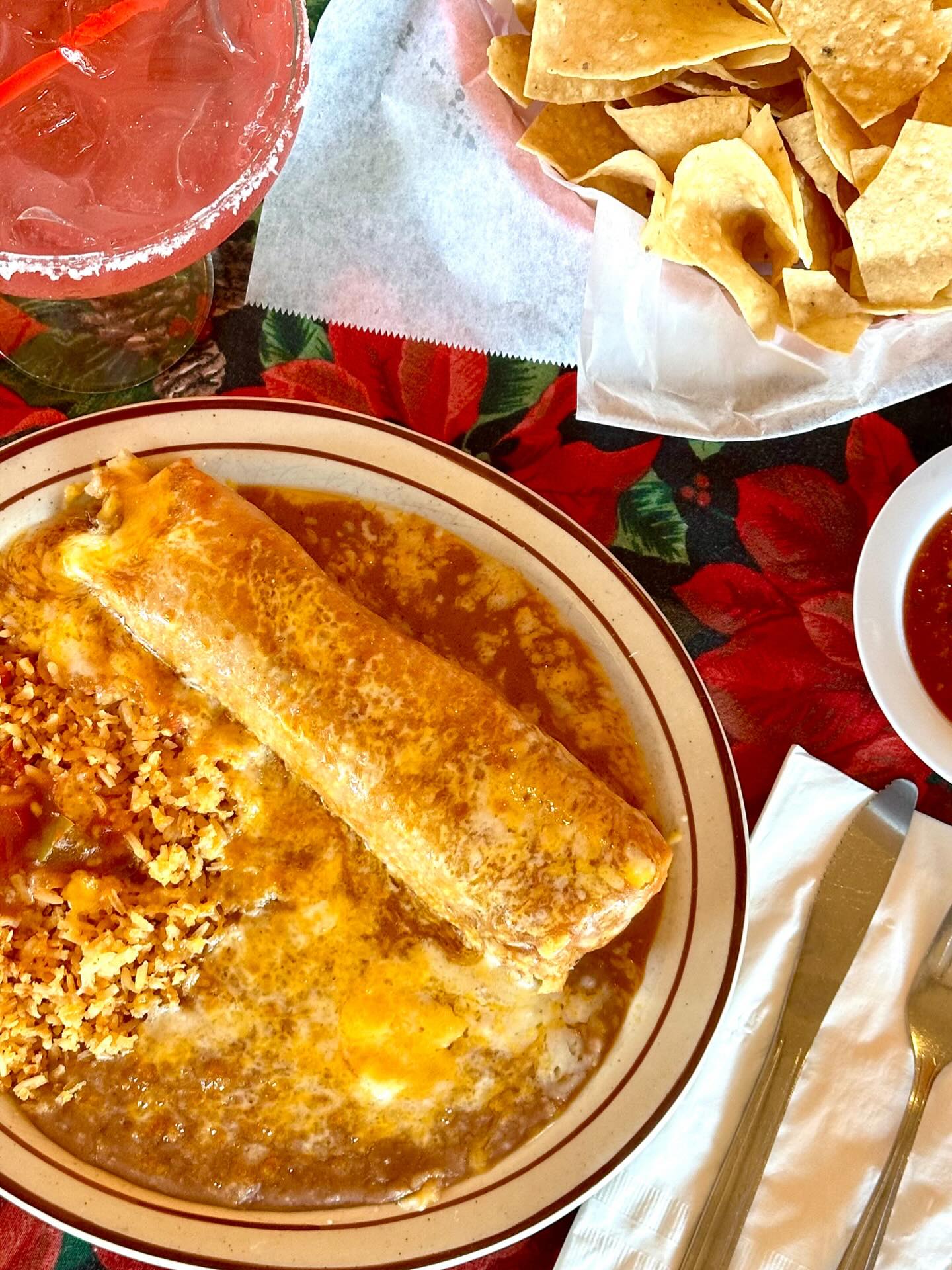 Plate with a smothered burrito topped in melted cheese and sauce, served with Mexican rice and refried beans, alongside tortilla chips and a pink margarita.