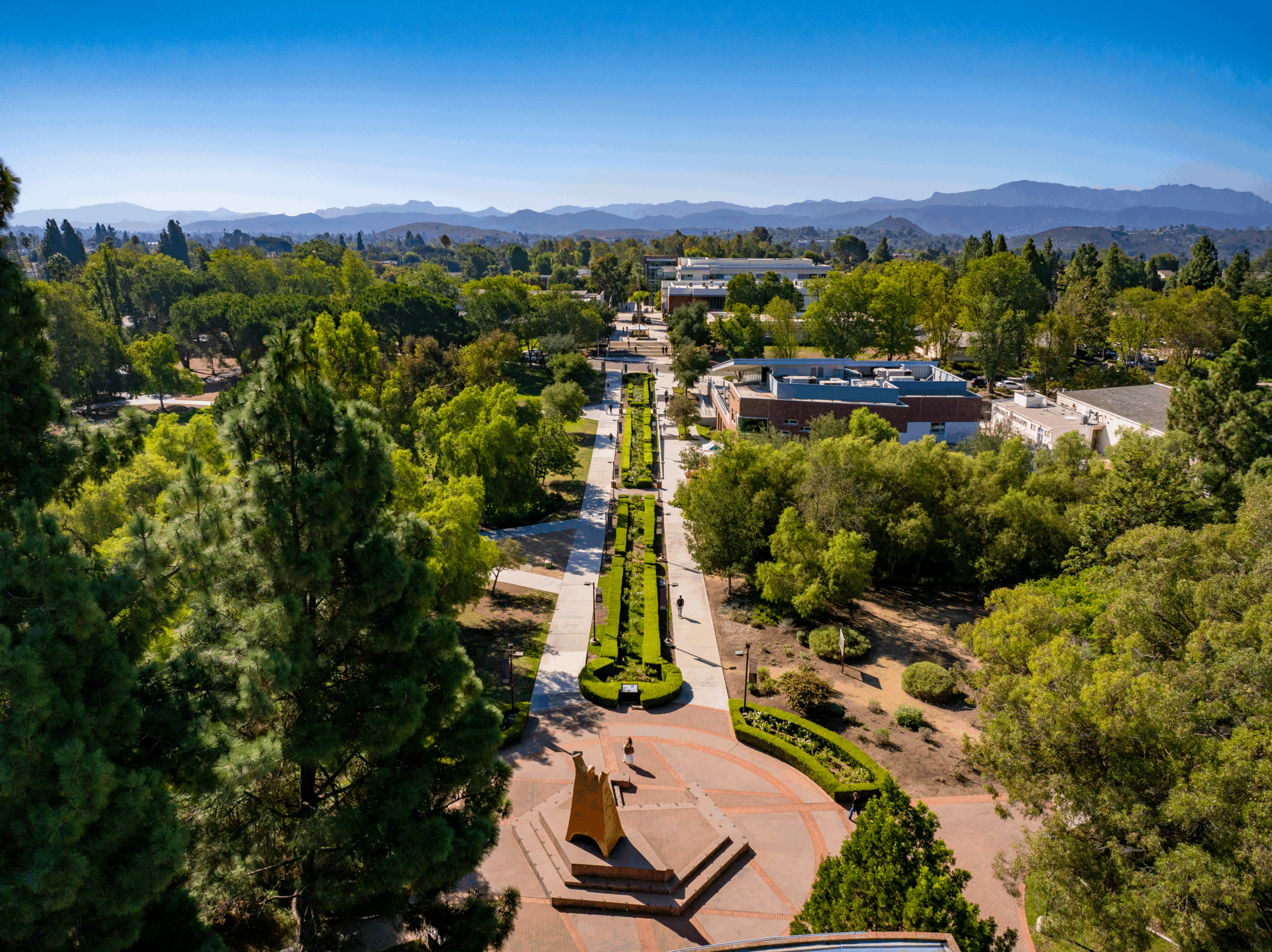 An aerial view of a tree-lined university campus with walkways, academic buildings, and mountains in the distance under a clear blue sky.