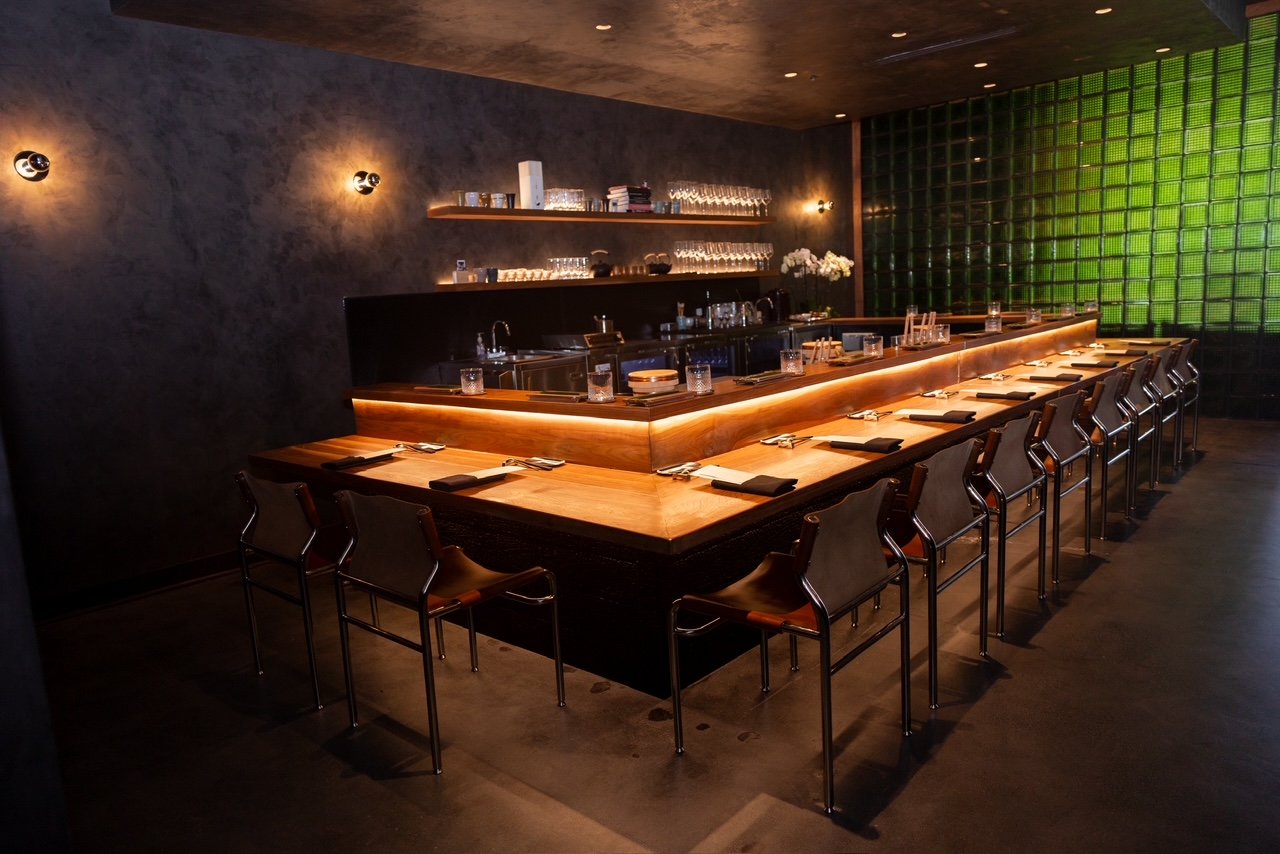Modern, dimly lit restaurant interior featuring a U-shaped wooden counter with bar seating, place settings arranged neatly, and a backlit green glass wall behind the bar.