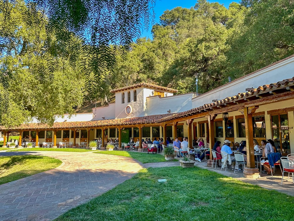 Outdoor courtyard at a Spanish-style winery with guests seated at patio tables beneath a covered walkway, surrounded by trees and hillside scenery.
