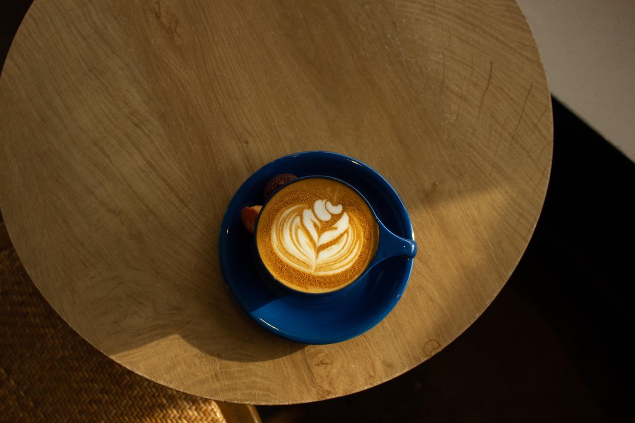 Blue cup of latte with leaf-shaped latte art on a small round wooden table.
