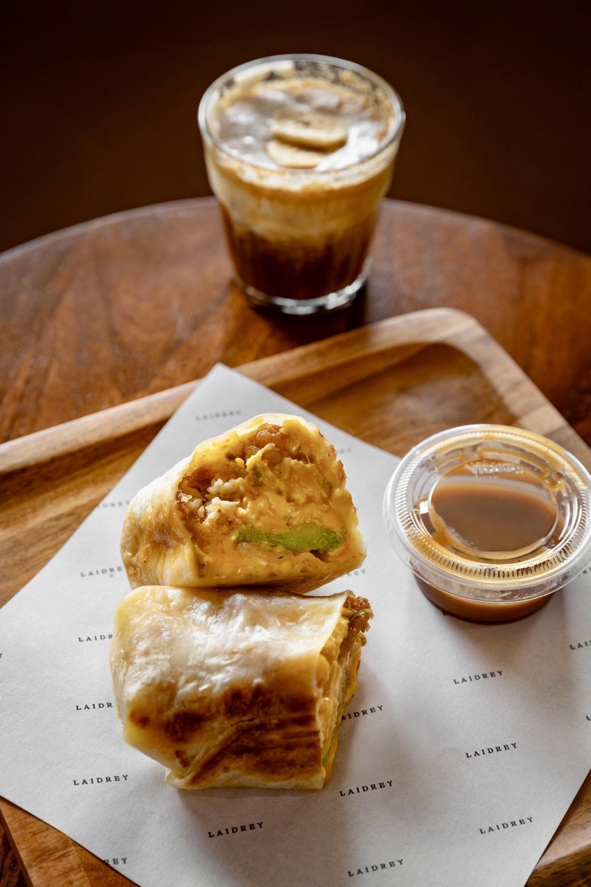 Breakfast burrito cut in half on a wooden tray with dipping sauce and an iced coffee in the background.