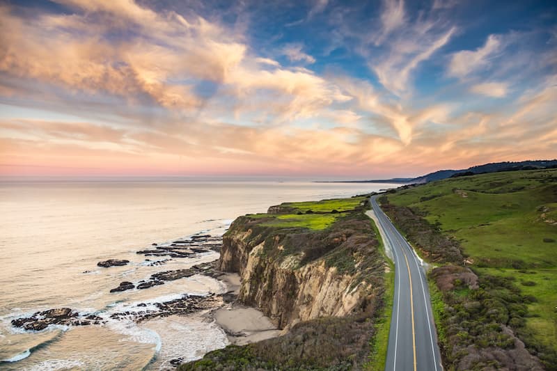 Scenic coastal highway winding along cliffs above the ocean at sunset with dramatic clouds.