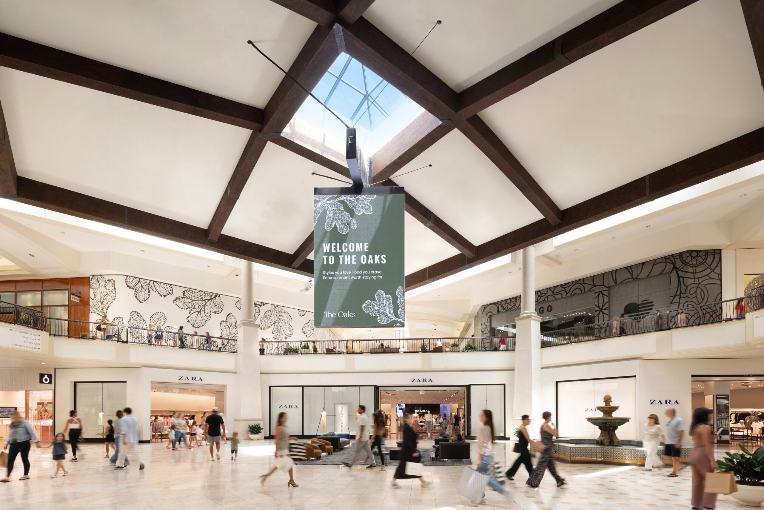 Open atrium inside a shopping mall with high ceilings, wood beams, a central hanging banner, and stores like Zara surrounding a busy floor.