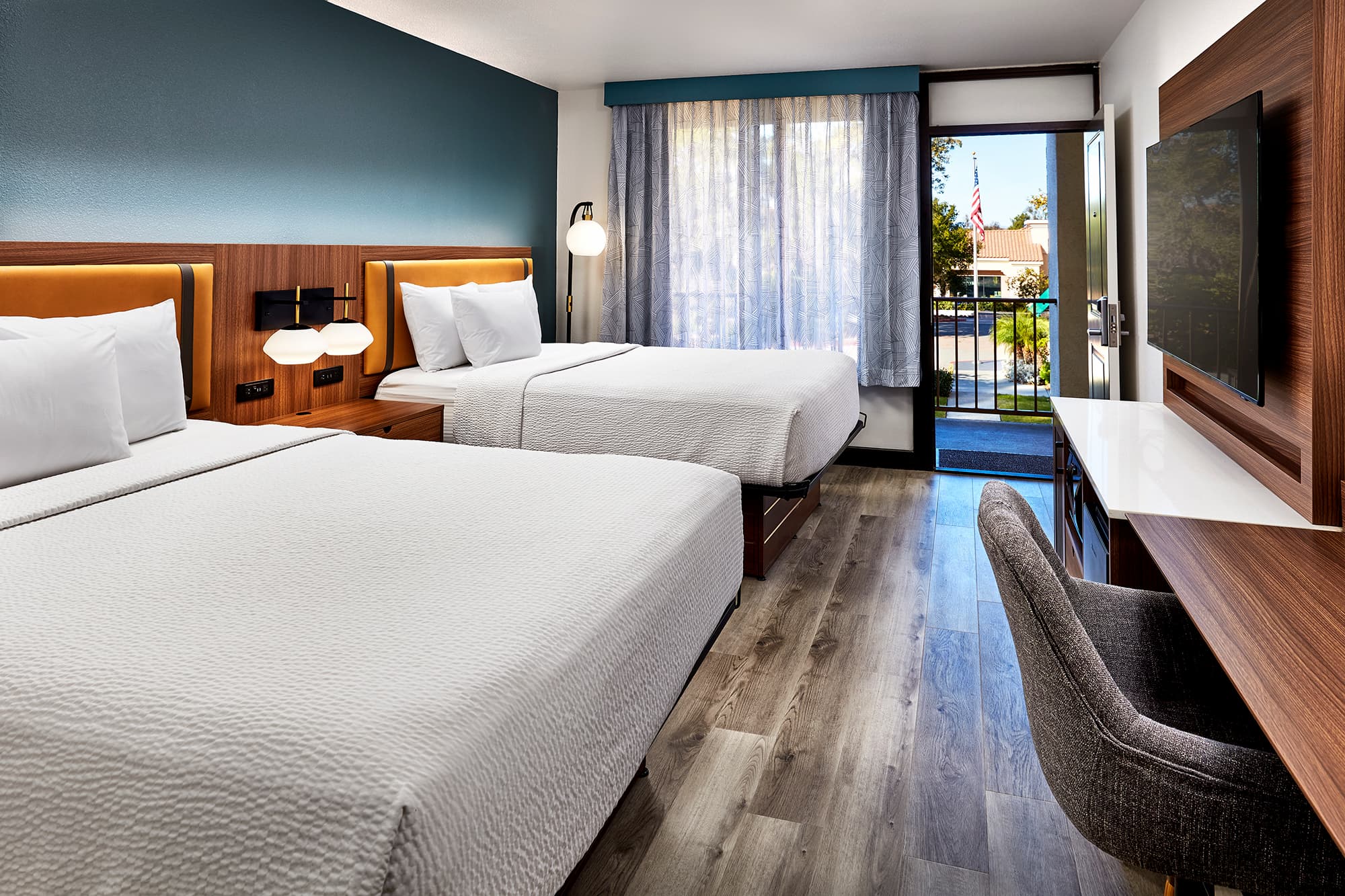 Contemporary hotel room with two queen beds, wood furnishings, a work desk, wall-mounted television, and an open balcony door letting in natural light.