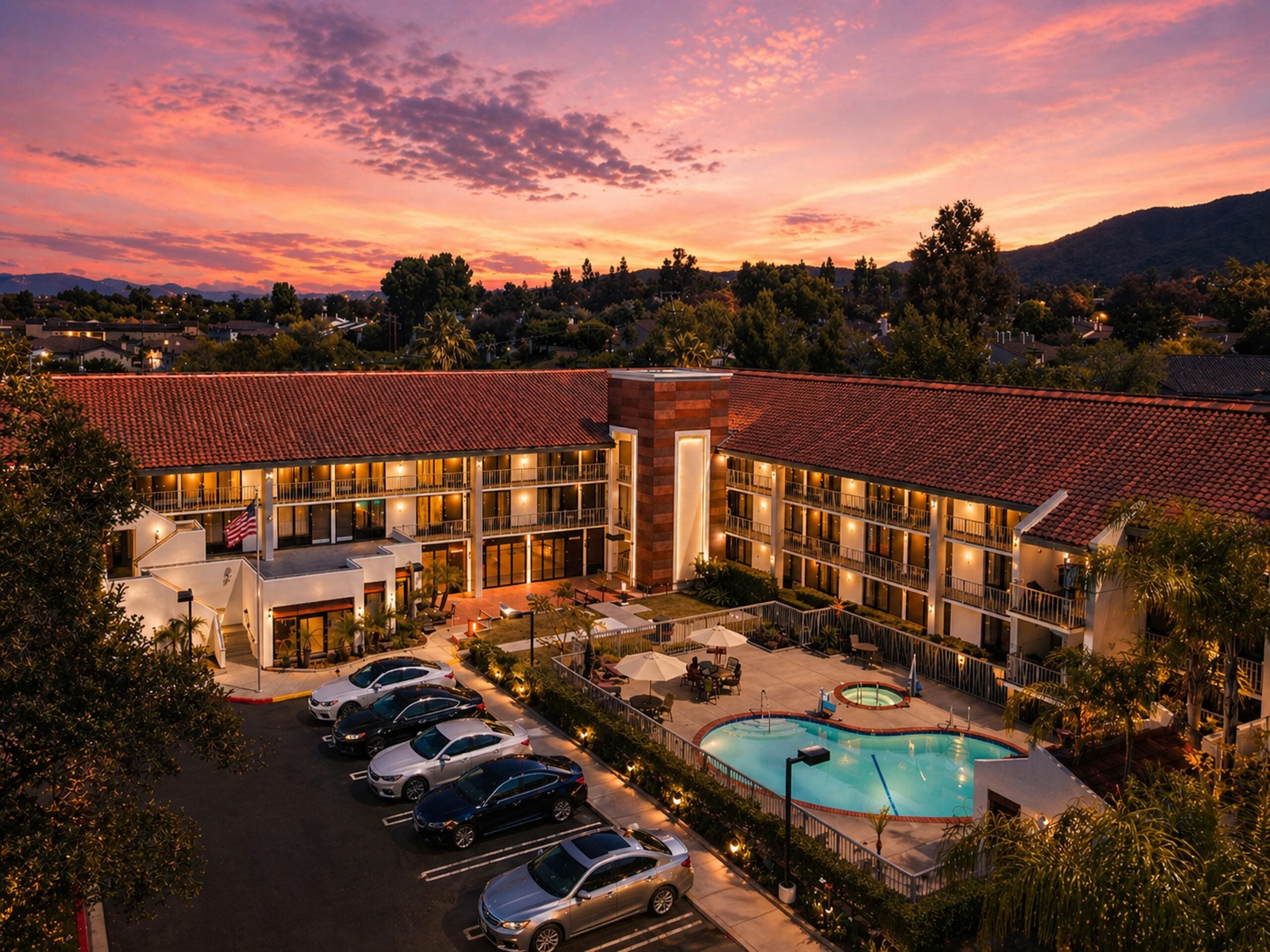 Aerial view of a La Quinta hotel at sunset featuring tiled roofs, illuminated guest rooms, a central courtyard pool and hot tub, parked cars, and mountains in the background under a colorful sky.
