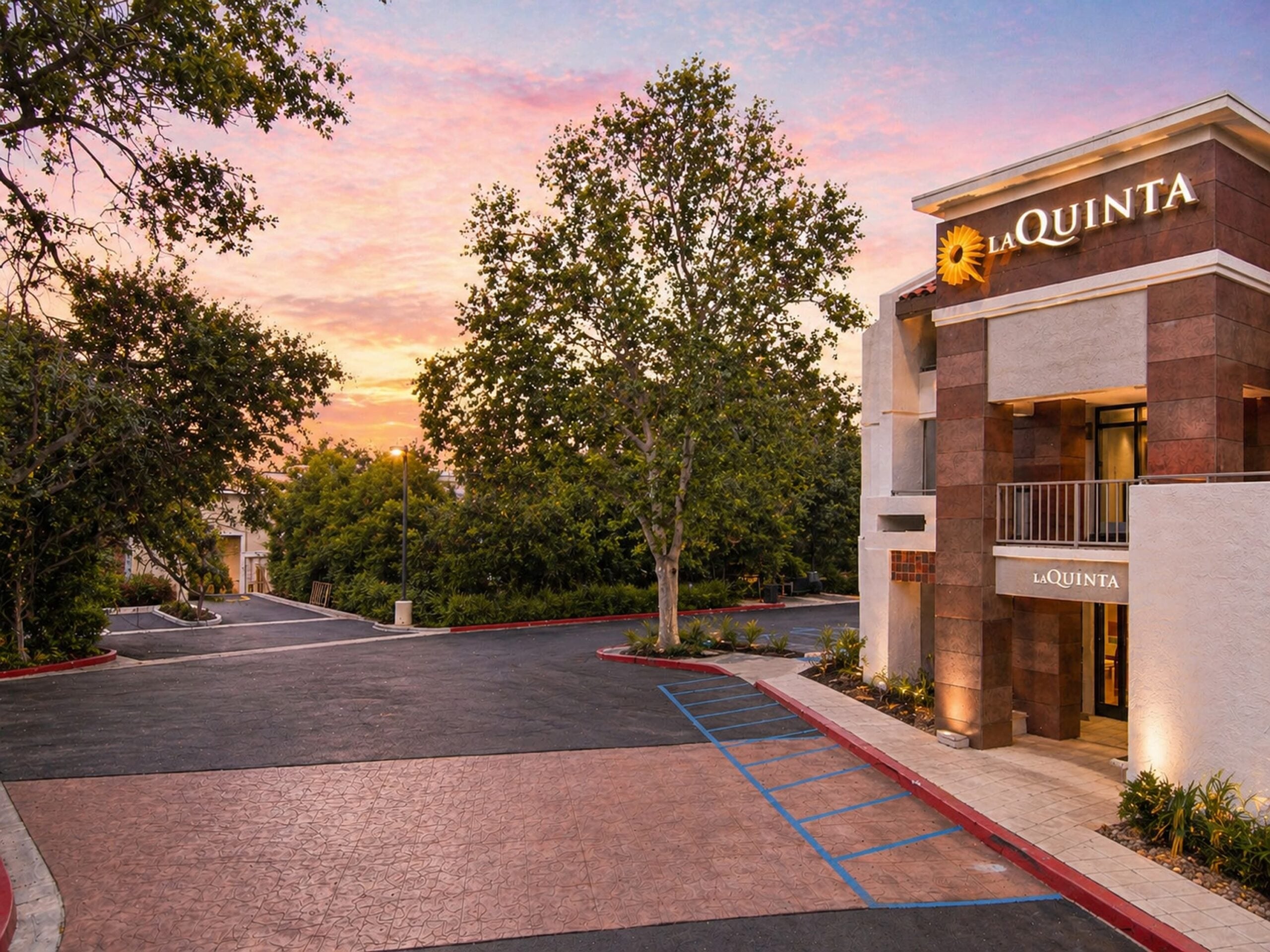 Front entrance of a La Quinta hotel surrounded by trees and landscaped grounds, with illuminated signage and a pink sunset sky overhead.