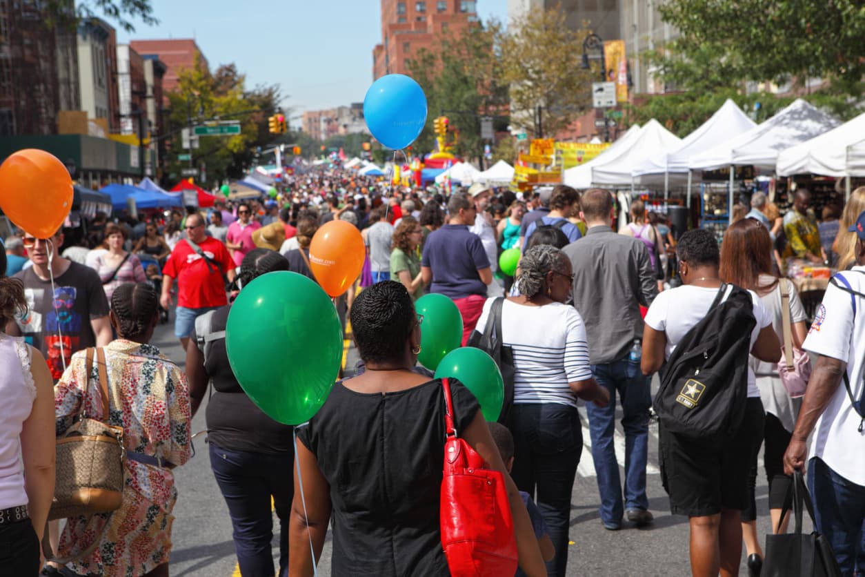 Large crowd of people walking through a busy outdoor street fair lined with vendor tents, colorful balloons, food stands, and market booths in a city setting.