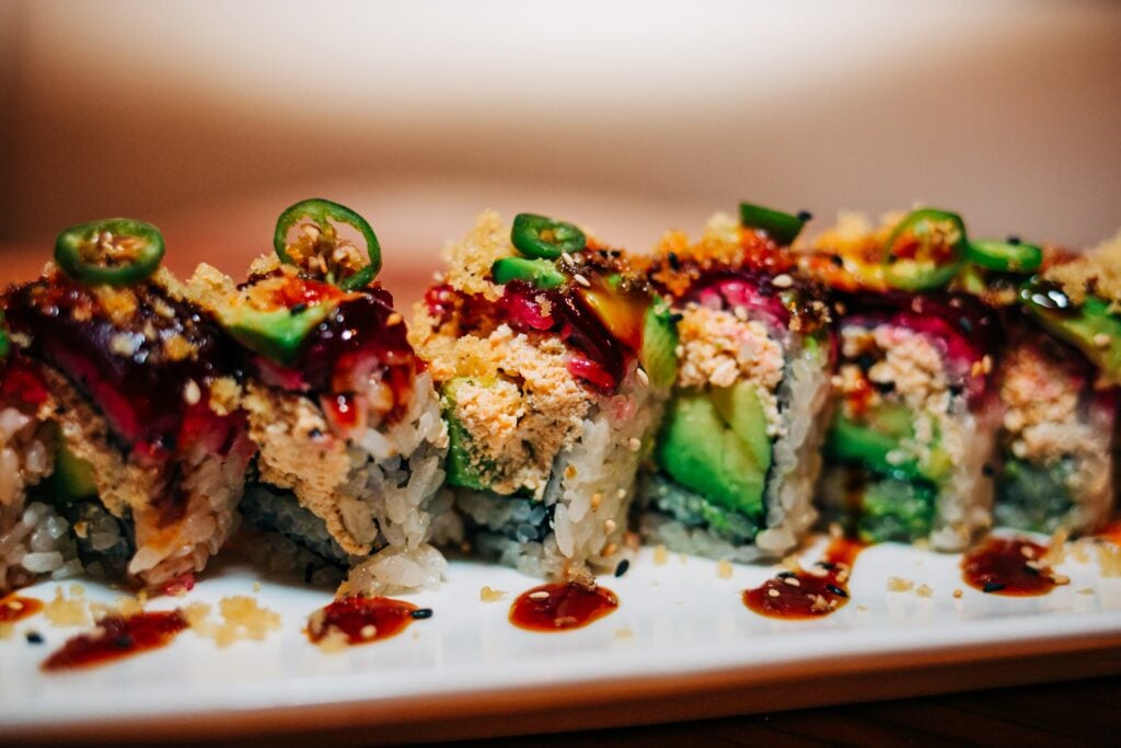 Colorful vegan sushi roll filled with avocado and topped with plant-based crumble, sliced jalapeños, sesame seeds, and sweet sauce drizzles on a white plate.