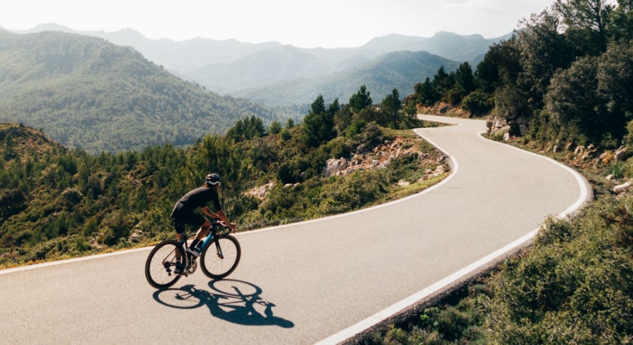Cyclist rides along a winding mountain road surrounded by lush green hills and distant peaks under a partly cloudy sky in Conejo Valley.