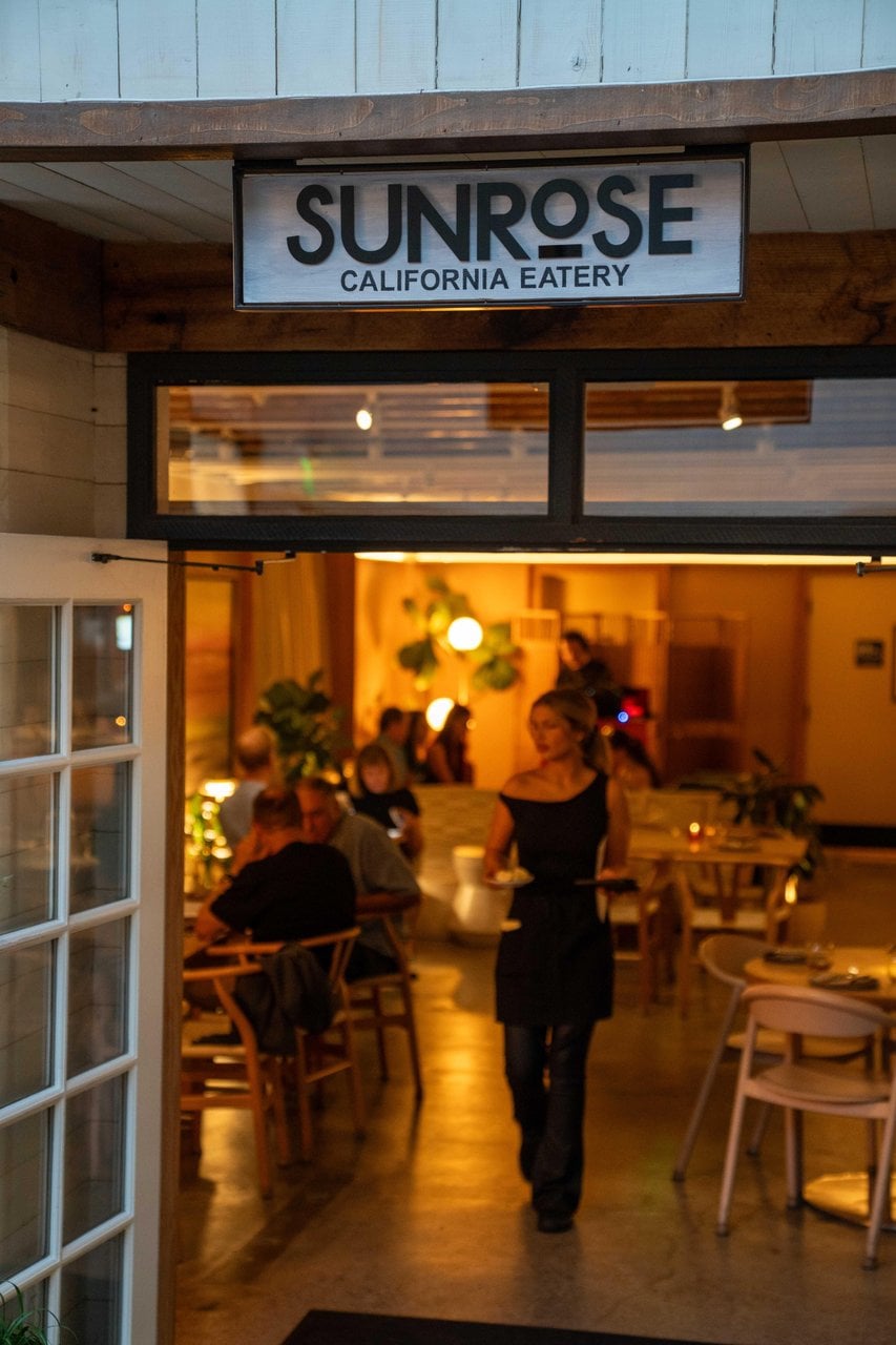 Evening view of the entrance to Sunrose California Eatery with diners enjoying the warmly lit dining room.