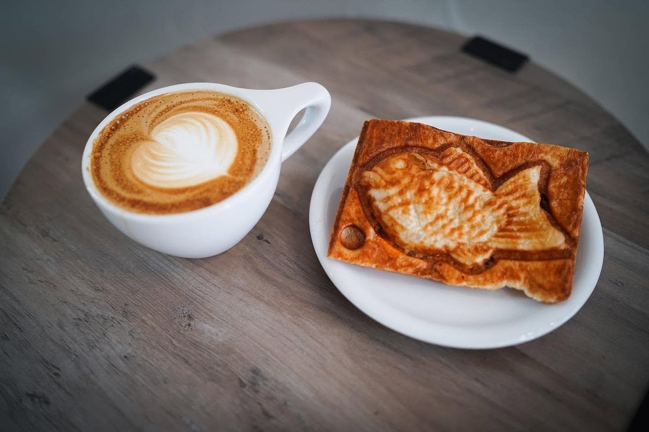 Cup of coffee with heart-shaped latte art beside a fish-shaped pastry served on a white plate.