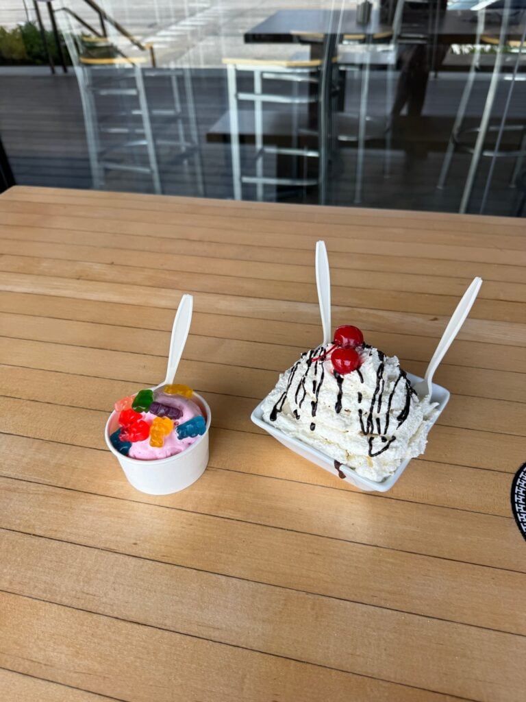 Two desserts on a wooden table: a small cup of pink ice cream topped with gummy bears and a larger whipped-cream-covered dessert drizzled with chocolate and topped with cherries.