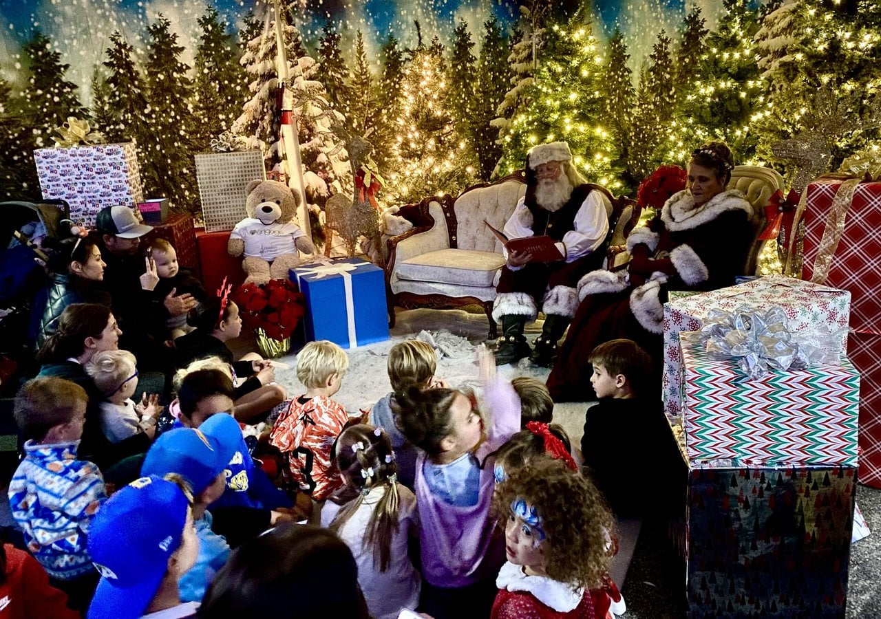 Children gathered around Santa and Mrs. Claus as Santa reads a book in a festive setting with Christmas trees and wrapped gifts.